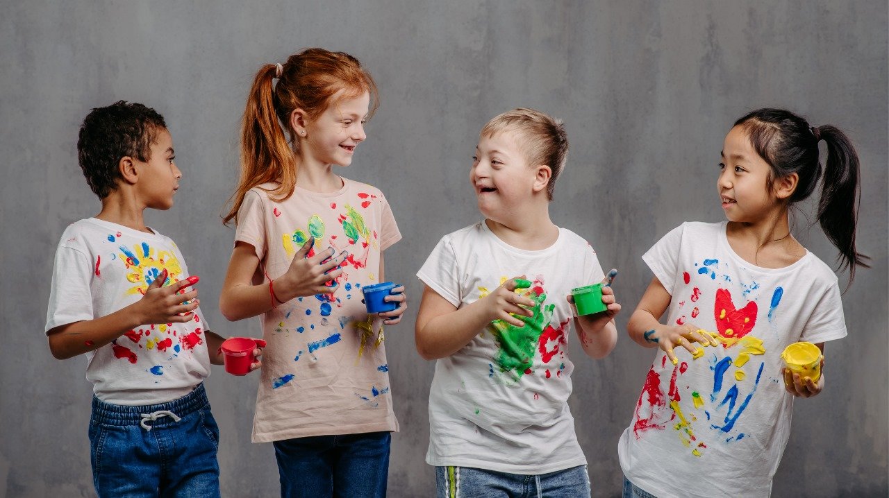 Três crianças sorrindo com as mãos e camisetas manchadas de tinta.